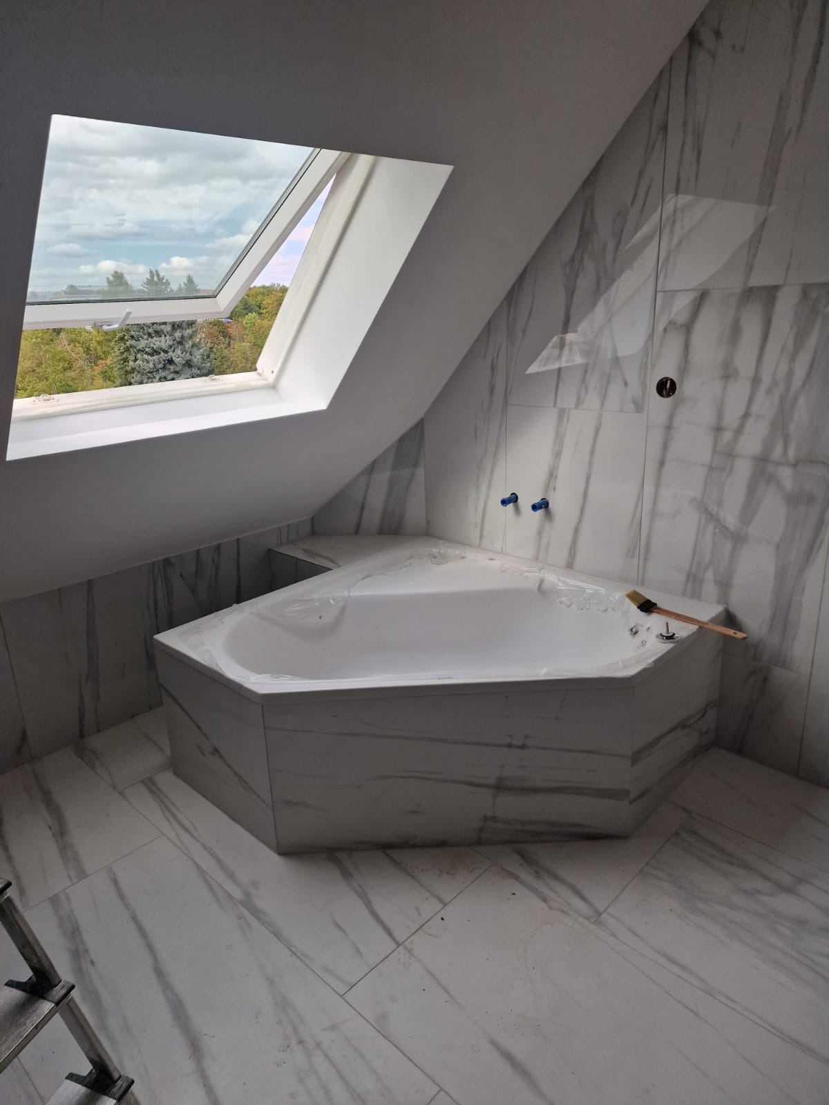 Wykończona łazienka na poddaszu z narożną wanną obudowaną marmurową płytką, widok na zewnątrz przez okno dachowe. Widoczna szczotka do fugowania.