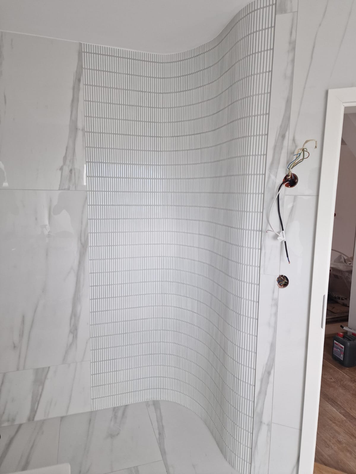 Wykończona, zakrzywiona ściana w łazience z białymi, marmurkowymi płytkami i mozaiką. Widoczne gniazdka elektryczne bez osprzętu.