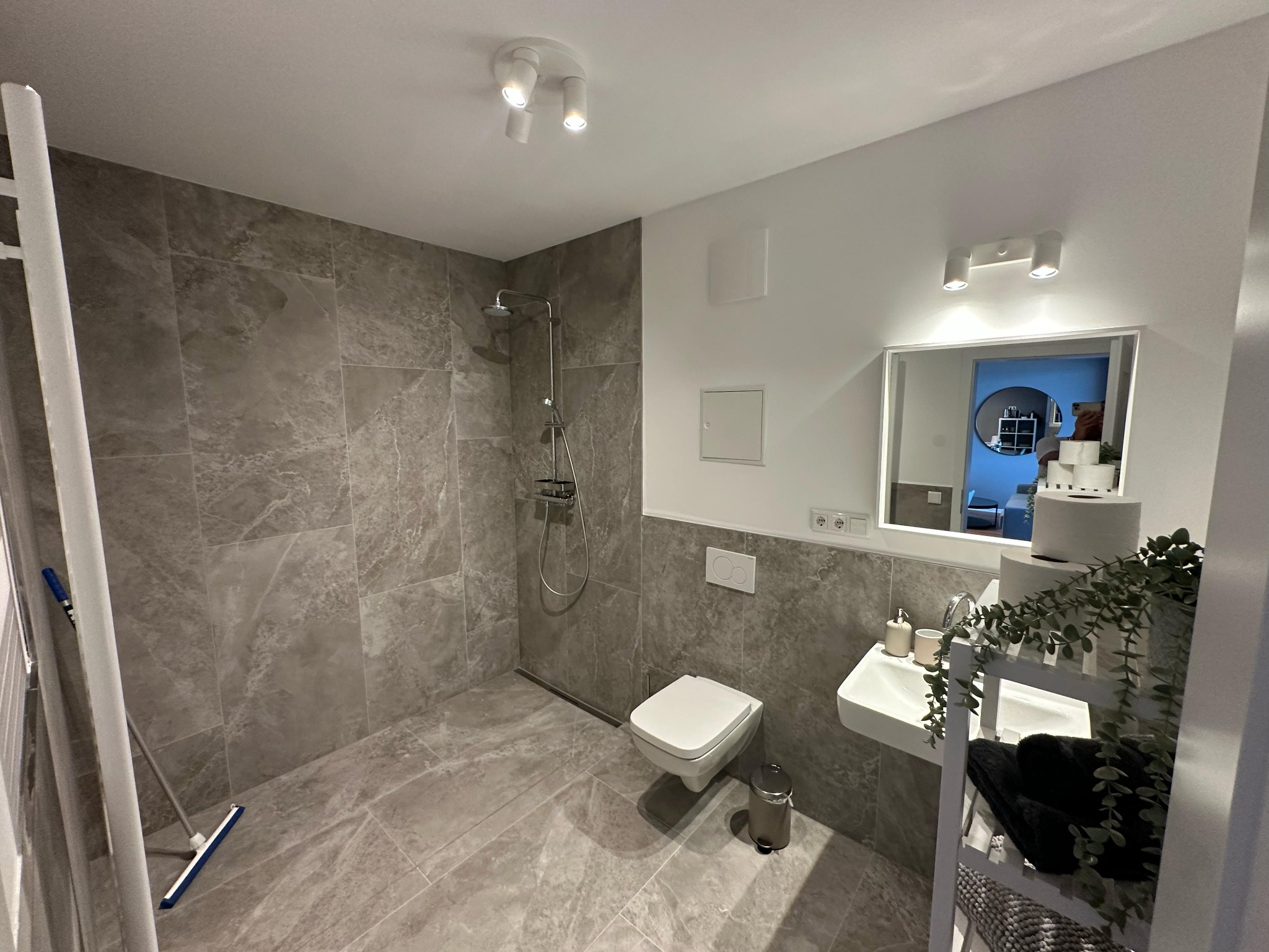 Nowoczesna łazienka w szarościach z kabiną prysznicową typu walk-in, umywalką i toaletą. Widoczne płytki imitujące marmur i oświetlenie punktowe.