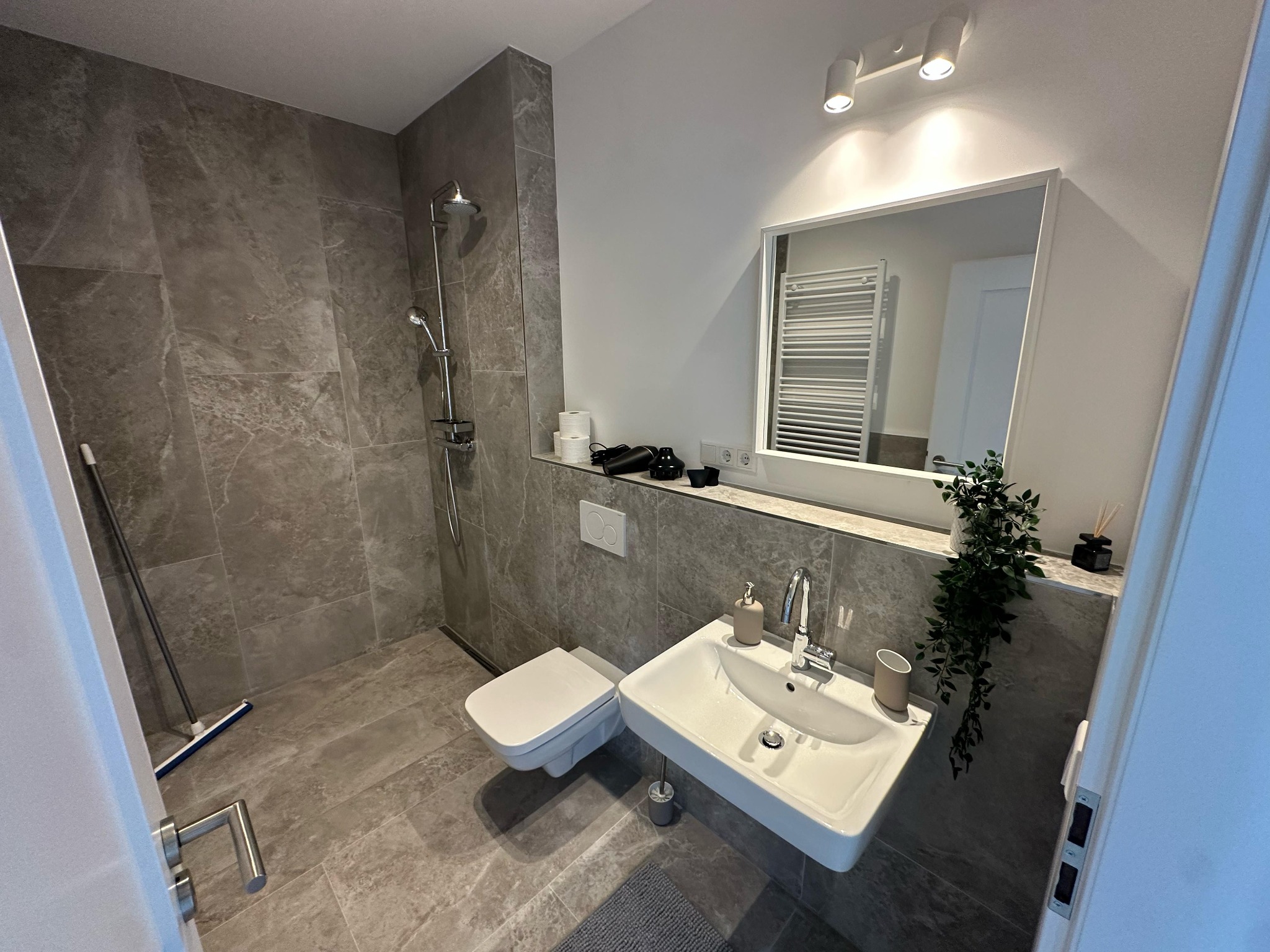 Nowoczesna łazienka z szarą, marmurkową glazurą, podwieszaną toaletą i umywalką. Widoczny prysznic, lustro i dekoracje.