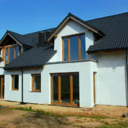 Nowoczesny dom jednorodzinny z brązowymi oknami i ciemną dachówką na tle błękitnego nieba w słoneczny dzień. Widok z boku na elewację.