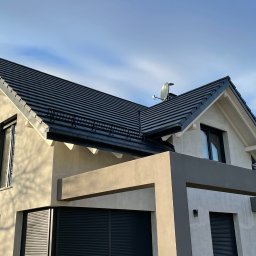 PPHU Pastenok Piotr Pasternok - Nowoczesny dom jednorodzinny z ciemną dachówką i jasną elewacją, widoczna antena satelitarna na dachu oraz cień drzewa na ścianie.