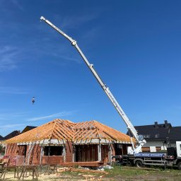PPHU Pastenok Piotr Pasternok - Budowa domu jednorodzinnego: wznoszenie drewnianej konstrukcji dachu przy użyciu dźwigu samochodowego, widoczne materiały budowlane i częściowo wymurowane ściany.
