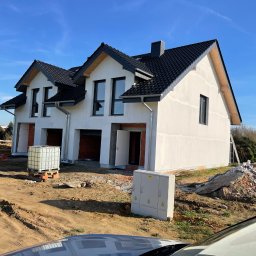 PPHU Pastenok Piotr Pasternok - Dwa bliźniacze domy w stanie surowym zamkniętym, z czarną dachówką i białymi ścianami, na tle błękitnego nieba. Widoczne materiały budowlane i skrzynka elektryczna przed budynkami.