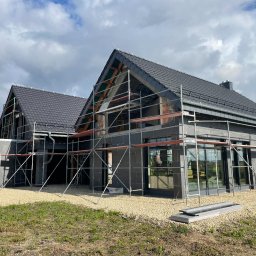 PPHU Pastenok Piotr Pasternok - Dwa domy w trakcie budowy z ciemnymi dachówkami, otoczone rusztowaniami i żwirem, widoczne elementy konstrukcji dachu.