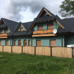 PPHU Pastenok Piotr Pasternok - Dom z ciemną dachówką karpiówką, drewnianymi balkonami i zieloną elewacją, ogrodzony płotem z jasnego drewna.
