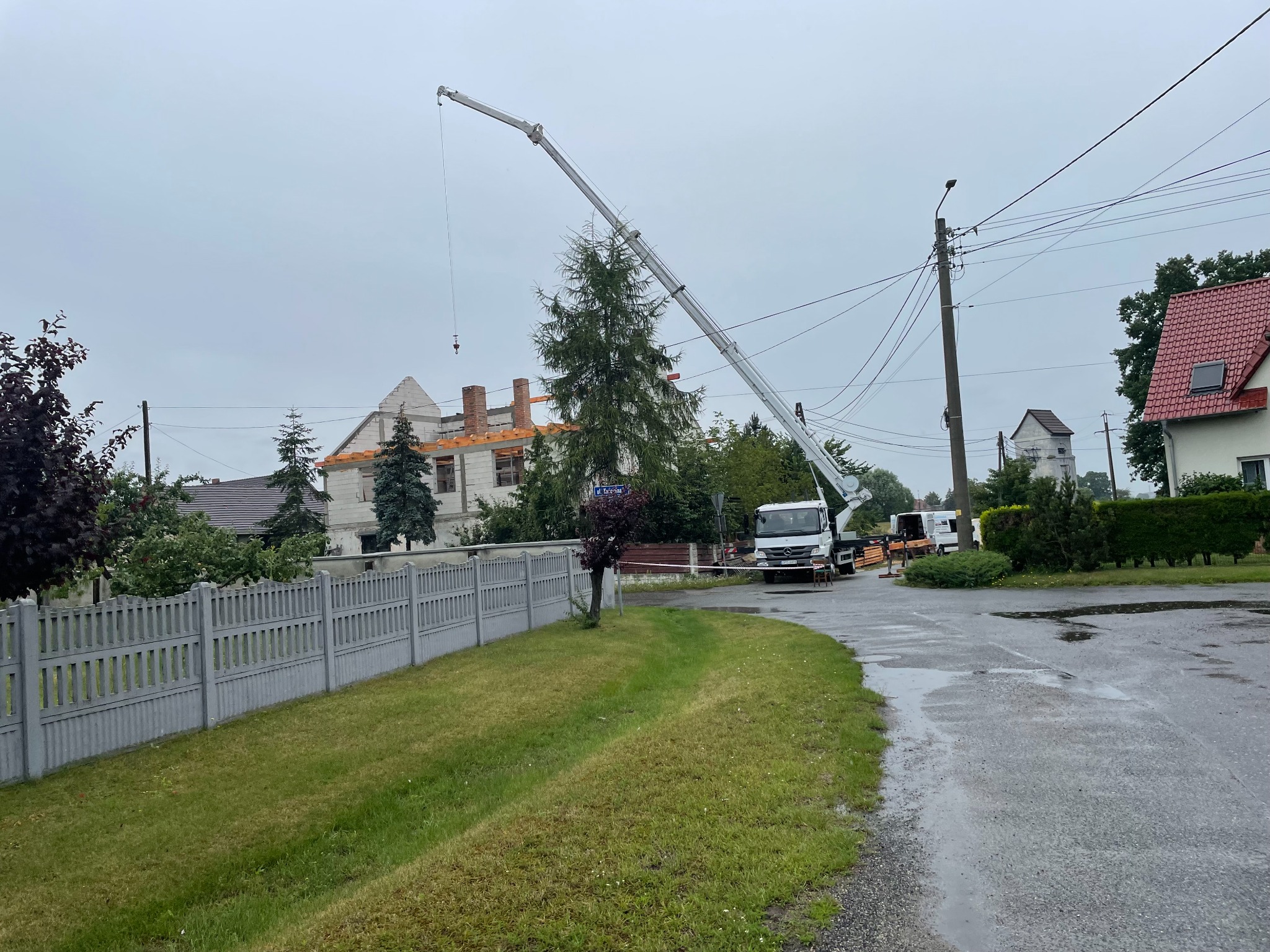Montaż więźby dachowej na budynku jednorodzinnym z użyciem dźwigu samochodowego na tle pochmurnego nieba i mokrej ulicy.