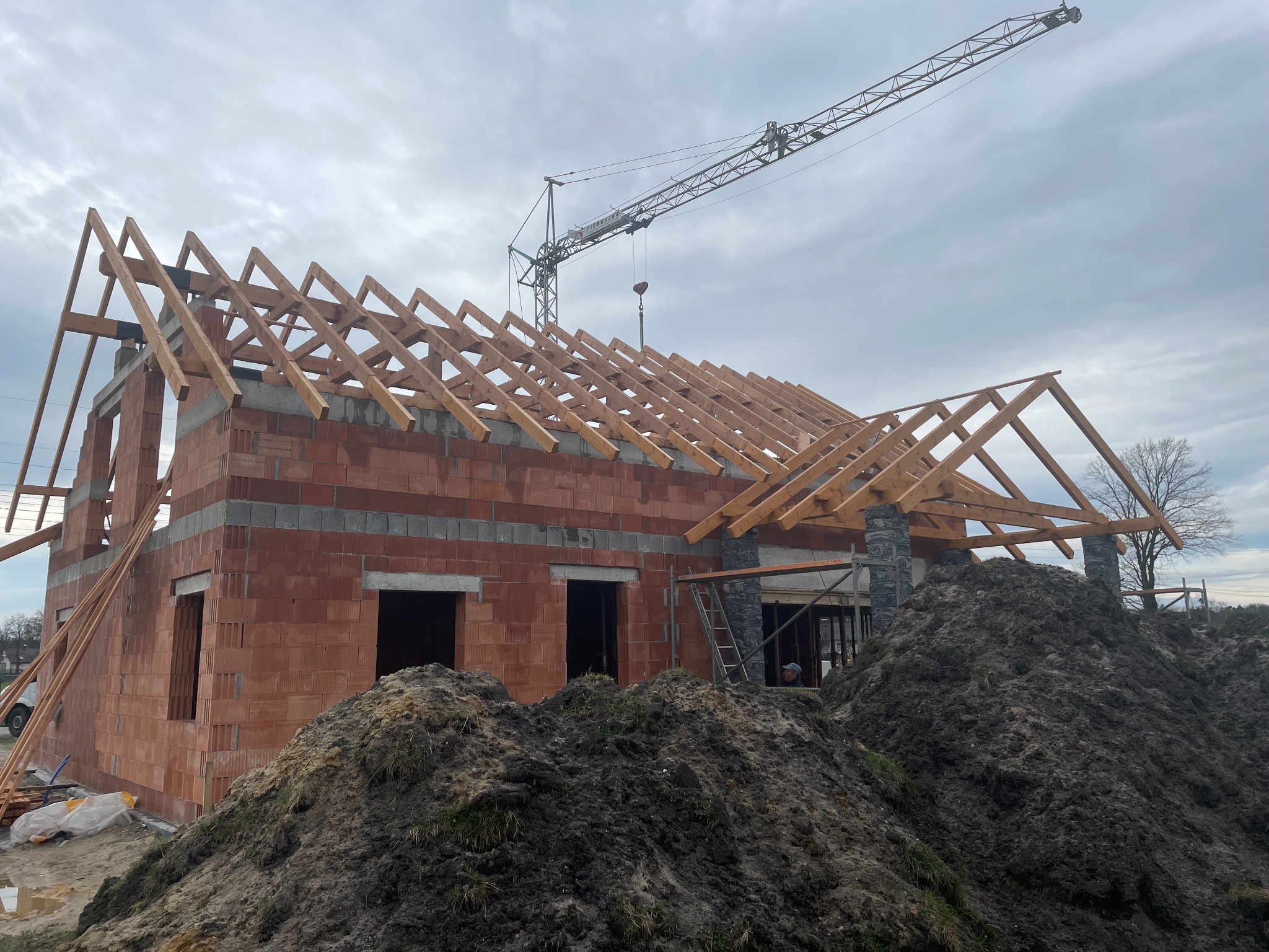 Budowa domu jednorodzinnego w trakcie wznoszenia konstrukcji drewnianej dachu, widoczny dźwig budowlany oraz sterta ziemi przed budynkiem.