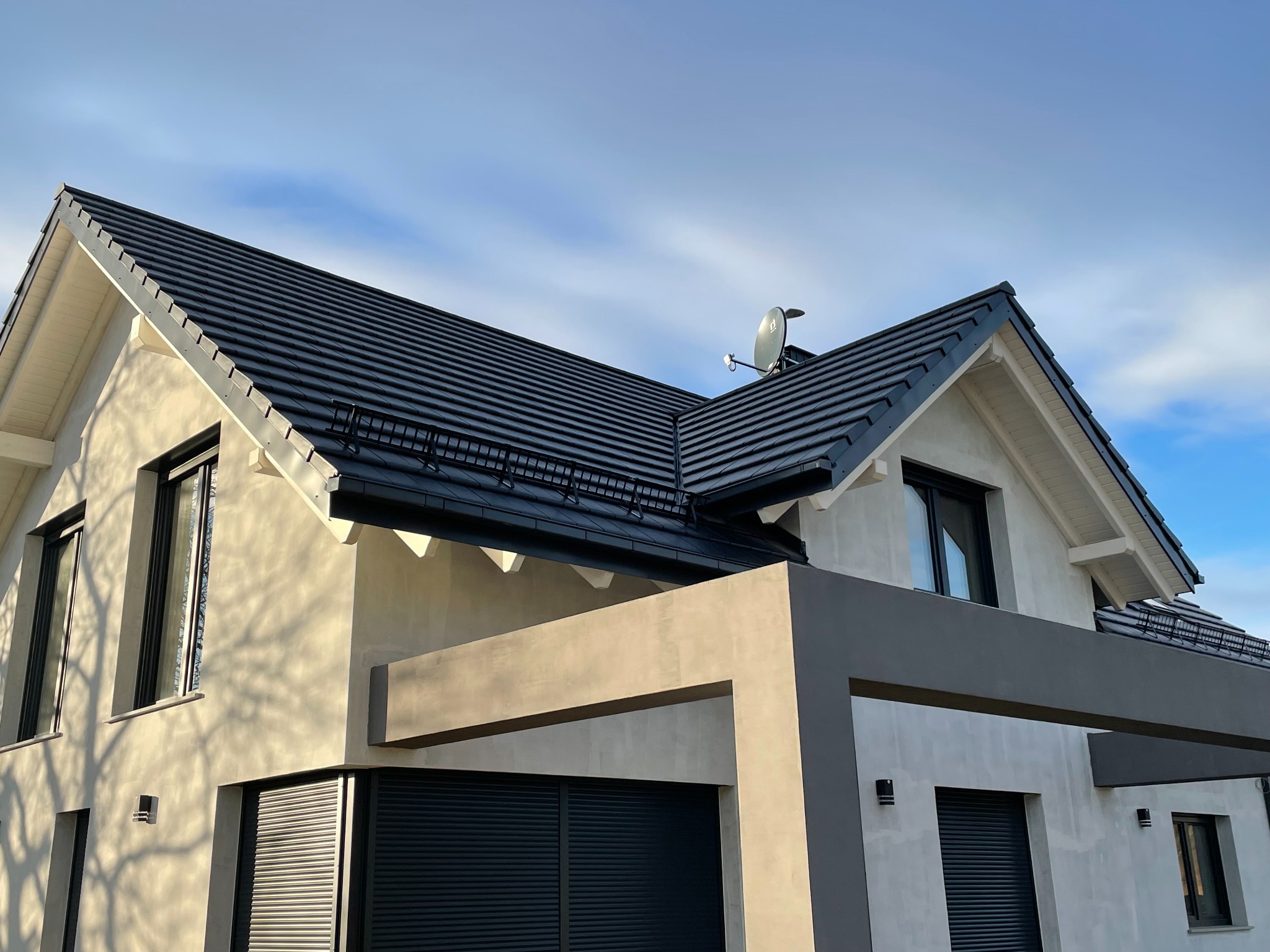 Nowoczesny dom jednorodzinny z ciemną dachówką i jasną elewacją, widoczna antena satelitarna na dachu oraz cień drzewa na ścianie.