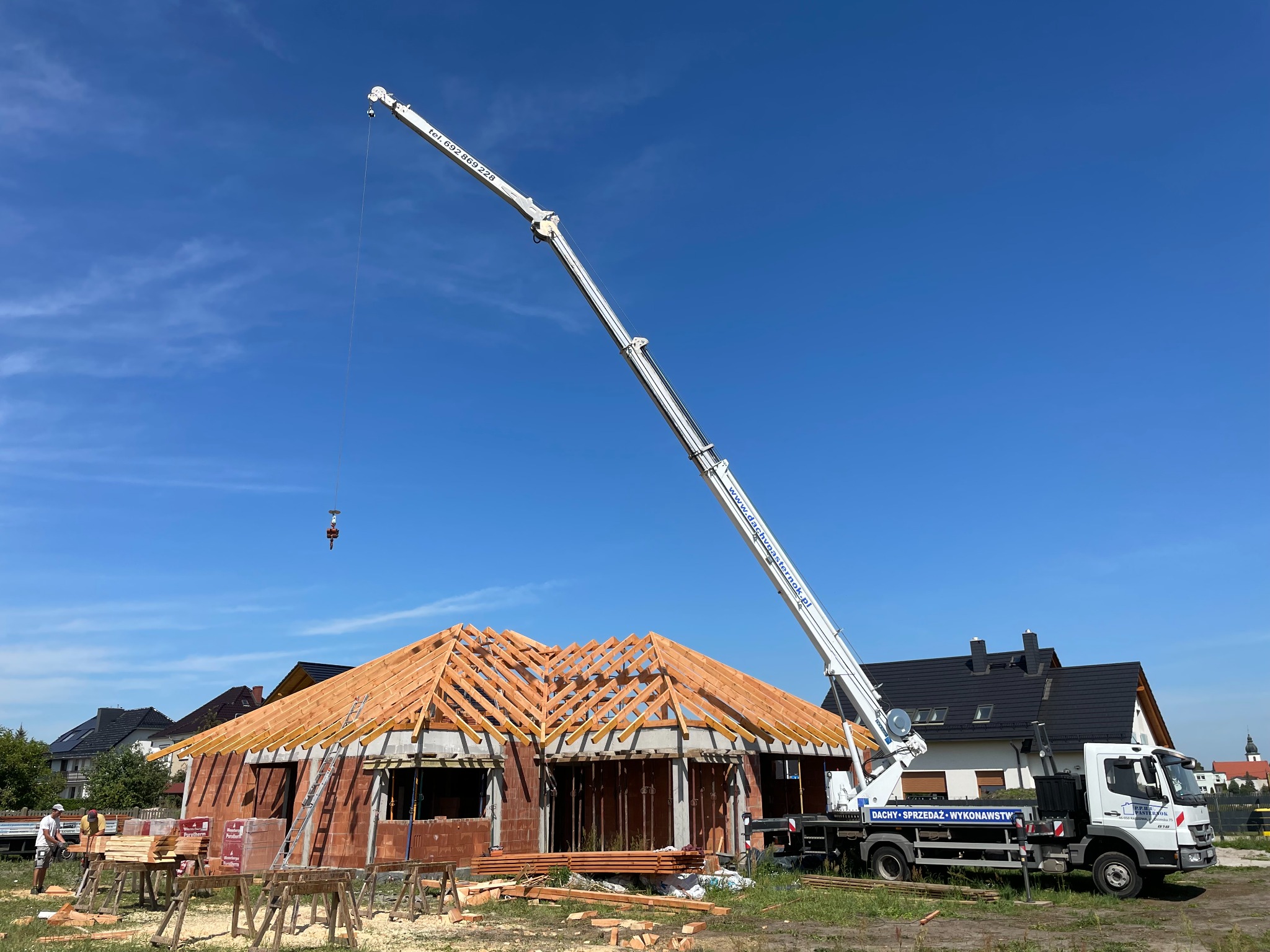 Budowa domu jednorodzinnego: wznoszenie drewnianej konstrukcji dachu przy użyciu dźwigu samochodowego, widoczne materiały budowlane i częściowo wymurowane ściany.