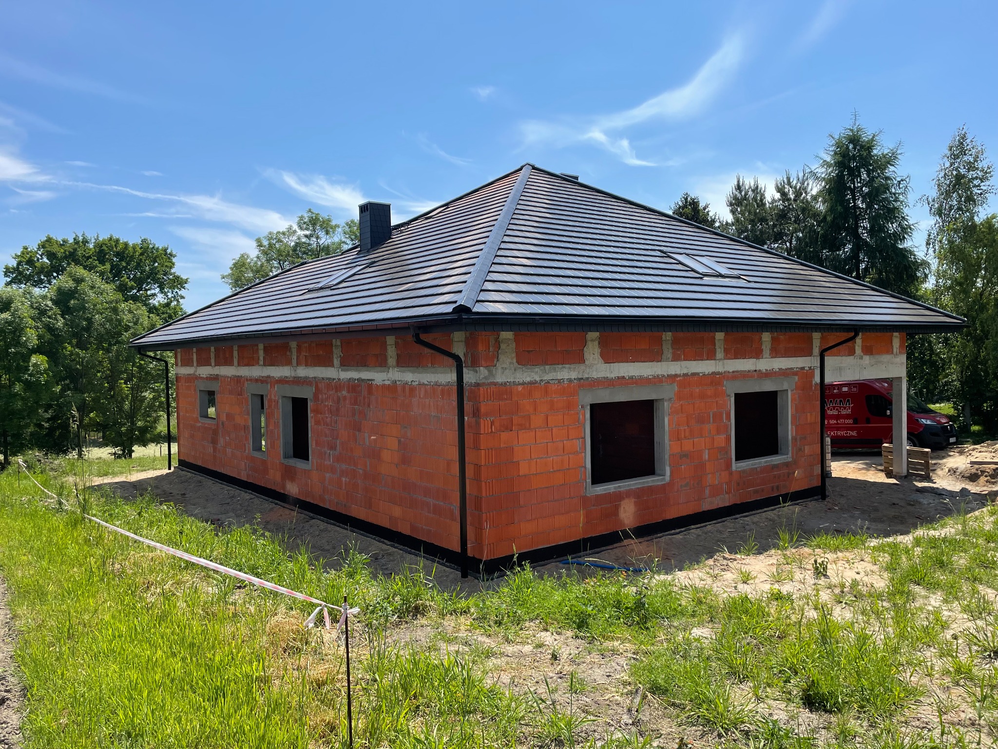 Surowy budynek jednorodzinny w trakcie budowy, z ciemną dachówką i czarnymi rynnami, widoczny fragment czerwonego samochodu dostawczego w tle.