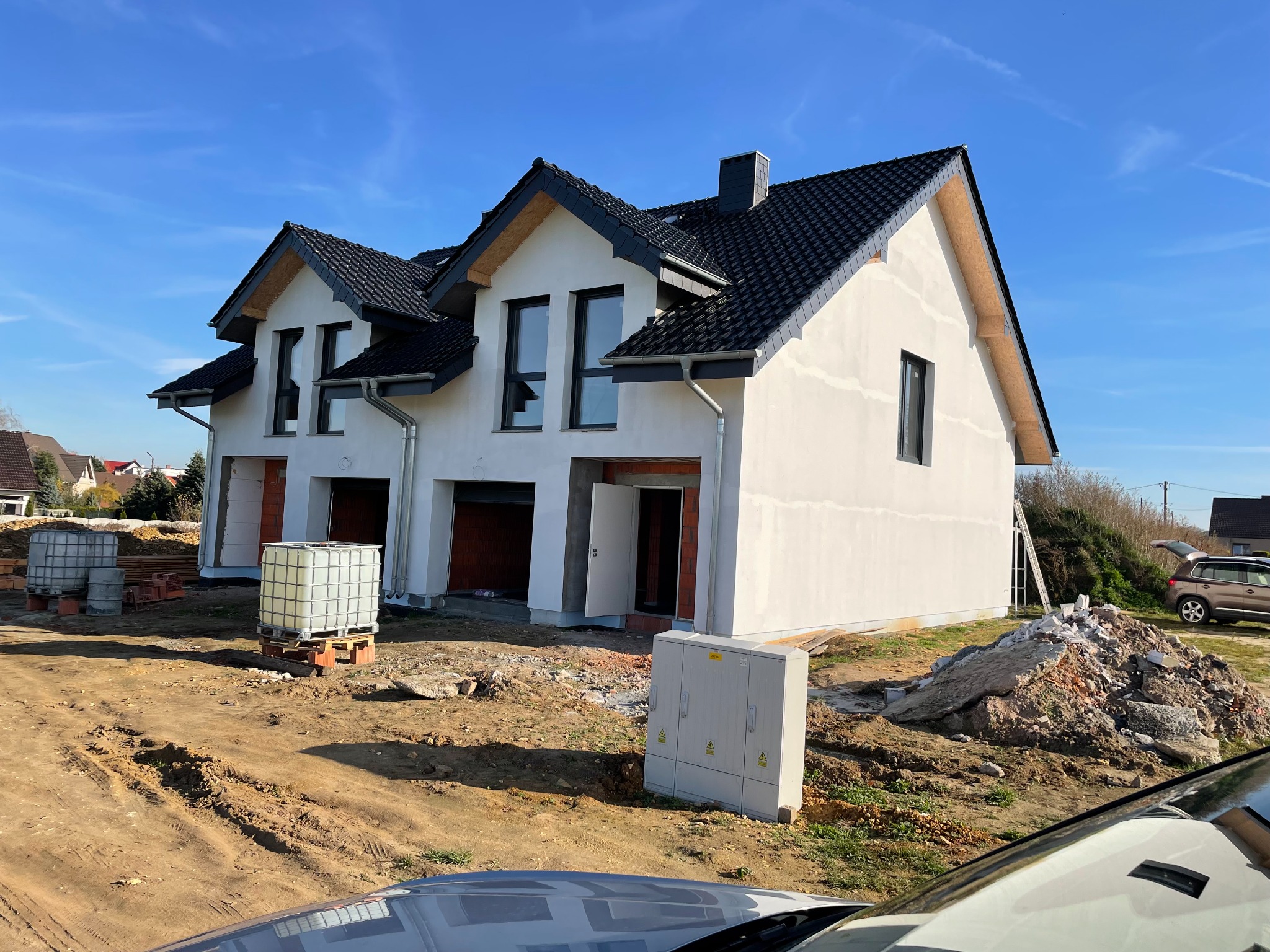 Dwa bliźniacze domy w stanie surowym zamkniętym, z czarną dachówką i białymi ścianami, na tle błękitnego nieba. Widoczne materiały budowlane i skrzynka elektryczna przed budynkami.