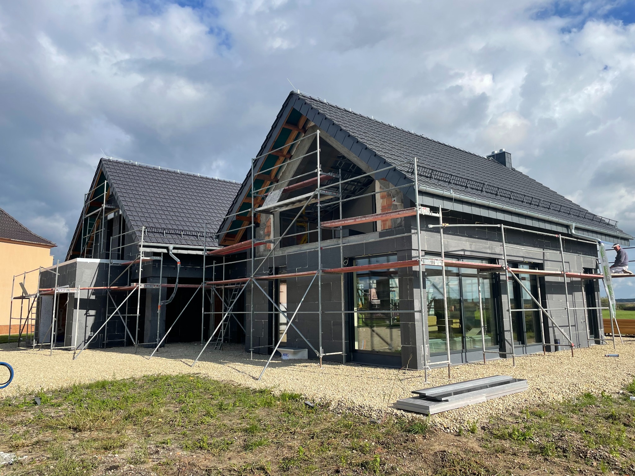 Dwa domy w trakcie budowy z ciemnymi dachówkami, otoczone rusztowaniami i żwirem, widoczne elementy konstrukcji dachu.