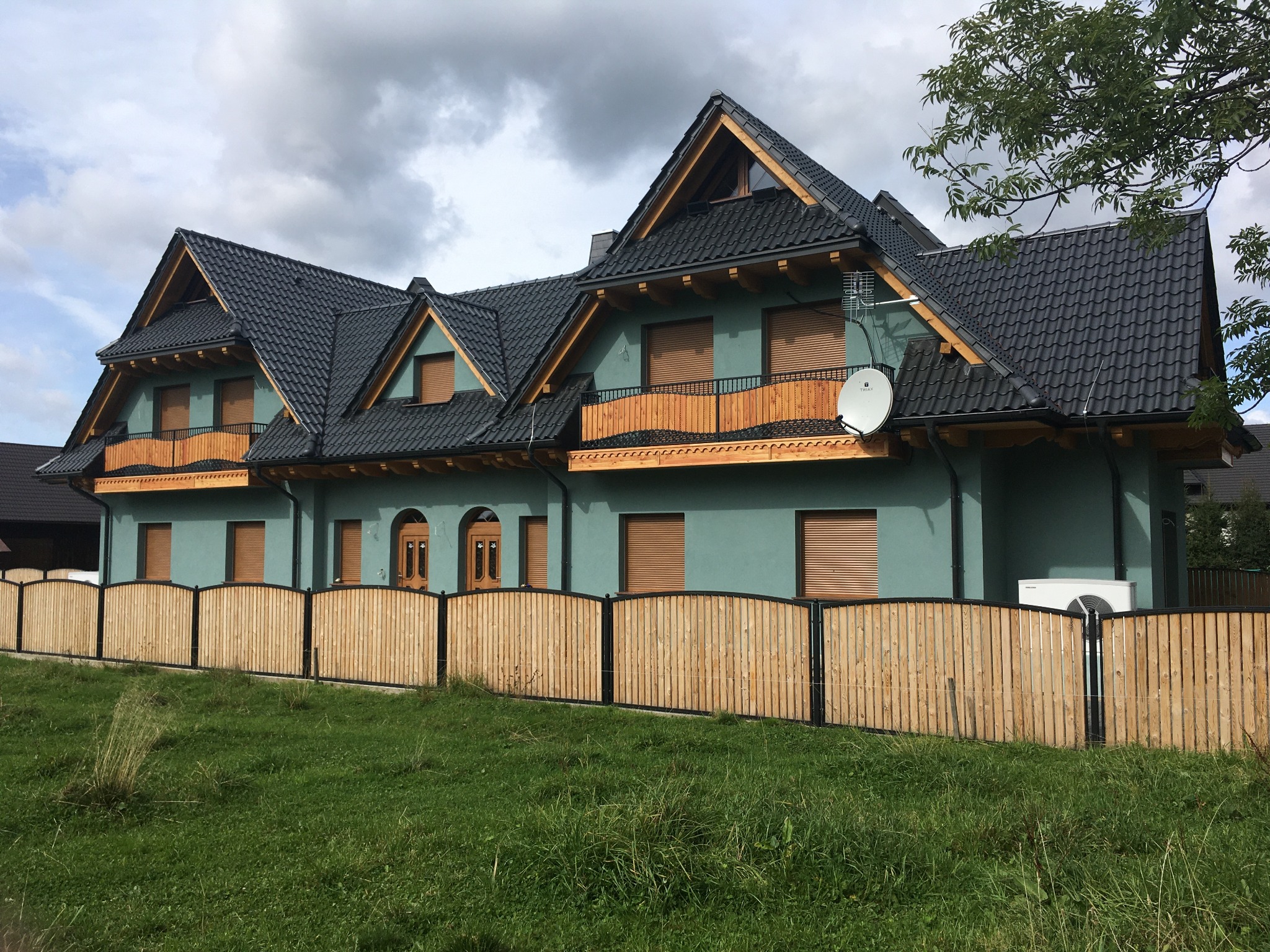 Dom z ciemną dachówką karpiówką, drewnianymi balkonami i zieloną elewacją, ogrodzony płotem z jasnego drewna.
