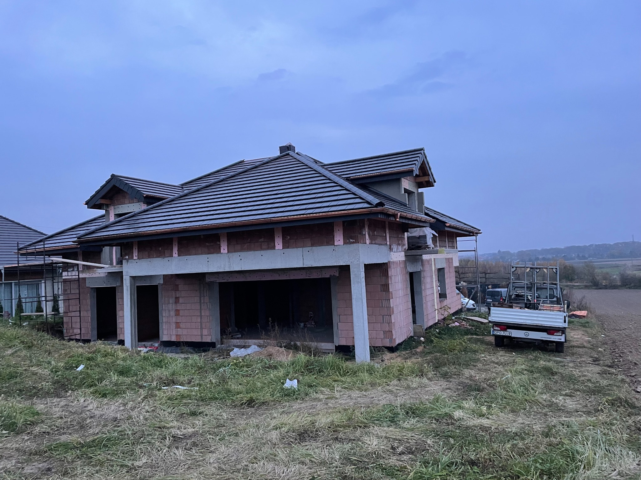 Niedokończony dom jednorodzinny z ciemną dachówką na częściowo ukończonym dachu, widoczna więźba dachowa, mur z czerwonej cegły i zaparkowany samochód dostawczy na trawiastym terenie.