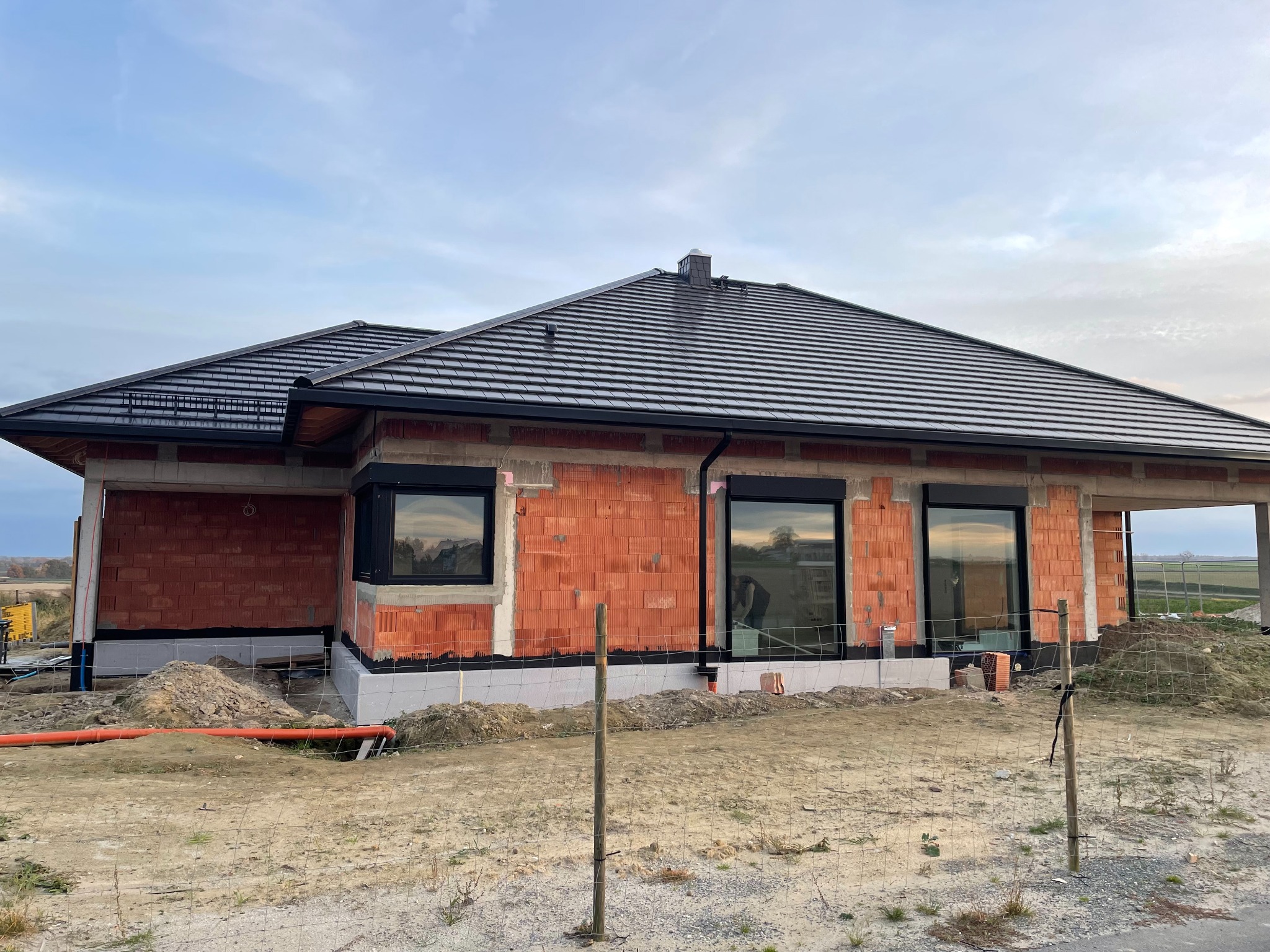 Budowa domu jednorodzinnego w trakcie realizacji, widoczna konstrukcja dachu z czarną dachówką, ściany z czerwonej cegły oraz teren wokół budynku.