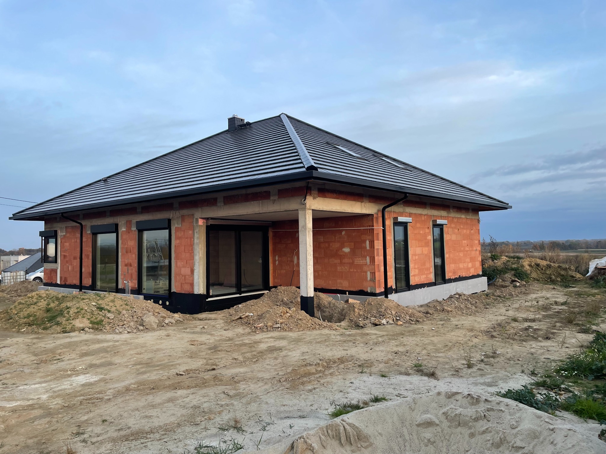 Surowy stan domu jednorodzinnego z czarną dachówką na skośnym dachu, widoczne okna z czarnymi ramami, ściany z czerwonej cegły i teren wokół w trakcie prac ziemnych.