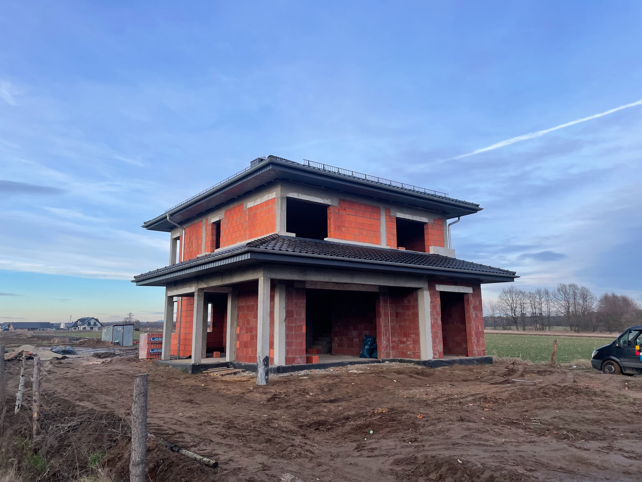 Niedokończony dwukondygnacyjny dom z czerwonej cegły z zamontowanymi szarymi rynnami, widoczne elementy konstrukcyjne dachu, teren budowy z błotem i sprzętem budowlanym.