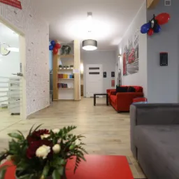 Jasne wnętrze szkoły językowej z recepcją w tle, czerwonymi sofami, drewnianą podłogą i ścianą pokrytą napisem 'NOW', ozdobione balonami i kwiatami.