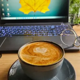 Filiżanka kawy latte na biurku z laptopem w tle, na ekranie żółty liść klonu. Okulary leżą obok filiżanki, sugerując przerwę w nauce.