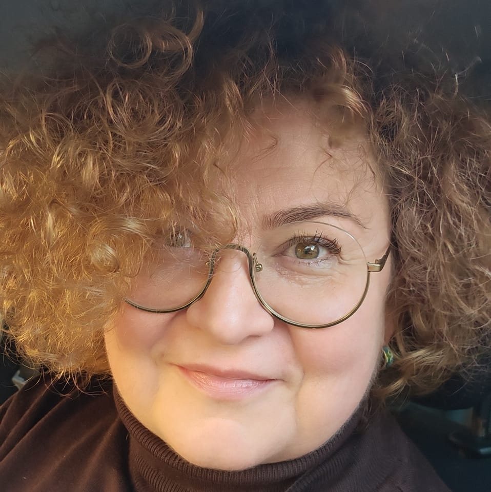 Portret kobiety z kręconymi włosami w okularach, ubrana w brązowy golf, uśmiechająca się do kamery.