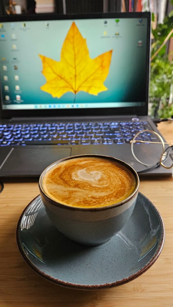 Filiżanka kawy latte na biurku z laptopem w tle, na ekranie żółty liść klonu. Okulary leżą obok filiżanki, sugerując przerwę w nauce.