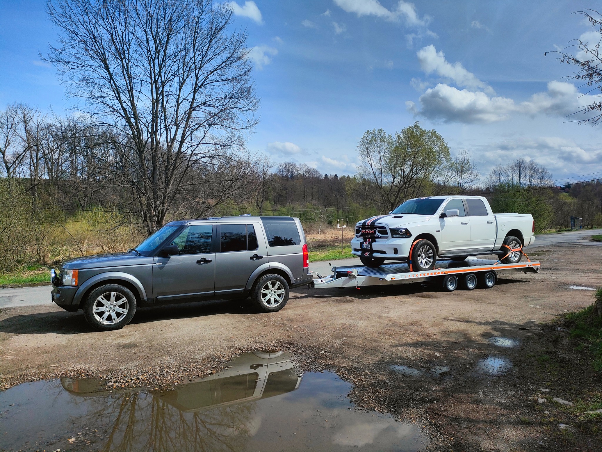 Szary SUV holuje na lawecie biały pickup z czarnymi pasami, widoczne pasy mocujące, tło stanowią drzewa i błękitne niebo z chmurami, na pierwszym planie kałuża odbijająca pojazdy.
