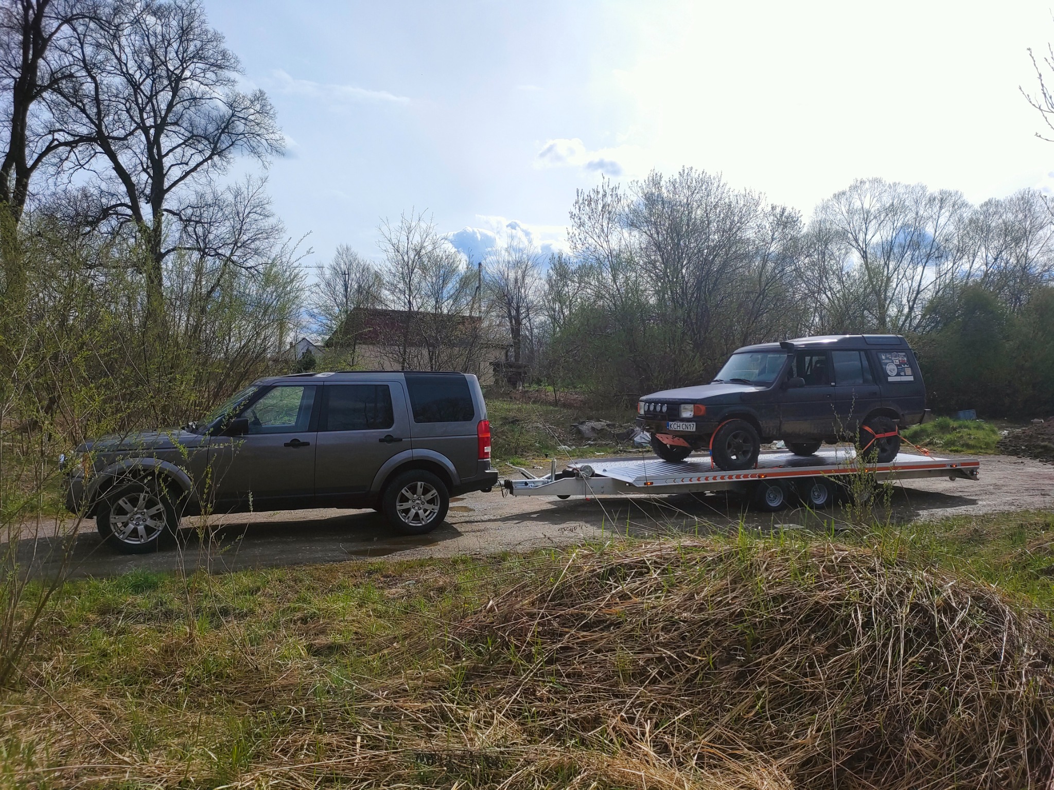 Szary samochód terenowy ciągnie na przyczepie lawetę z czarnym samochodem terenowym, widok z boku na tle drzew i nieba.