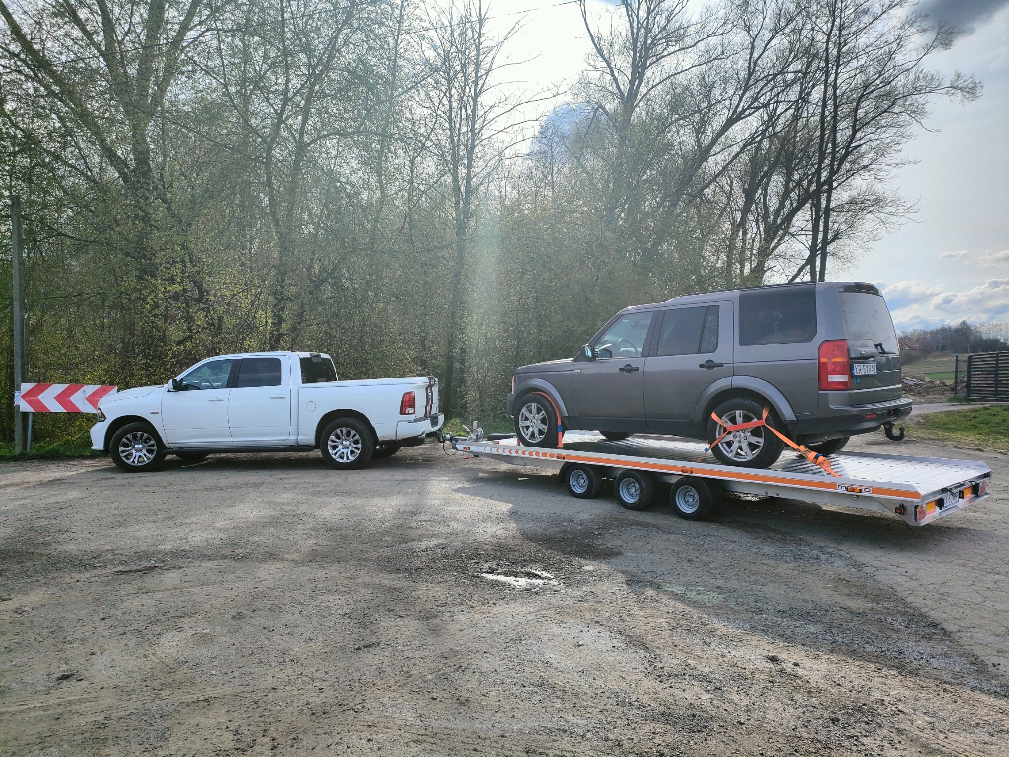 Biały pickup holuje szarą terenówkę na niskopodwoziowej przyczepie w słoneczny dzień, na ziemistej nawierzchni, z drzewami w tle.
