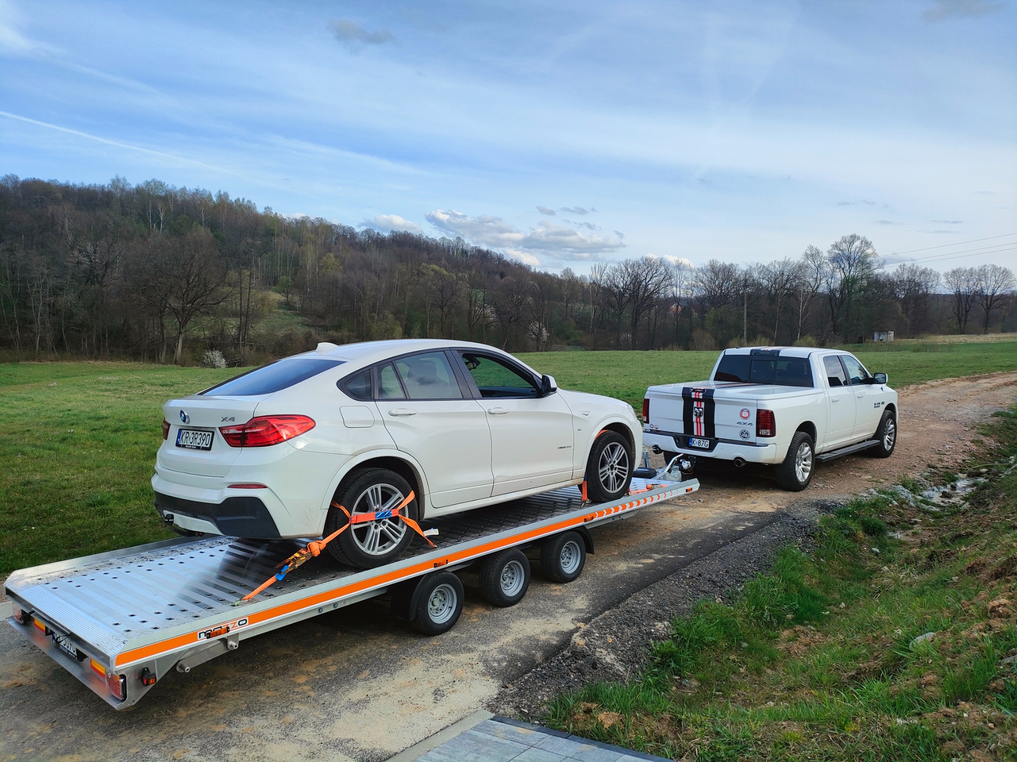 Biały samochód BMW X4 zabezpieczony pomarańczowymi pasami na srebrnej platformie lawety, ciągniętej przez białą ciężarówkę Dodge Ram na polnej drodze z widokiem na łąkę i las w tle.