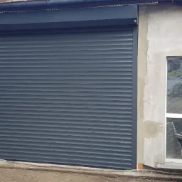 Zamknięta grafitowa roleta zewnętrzna na szarej ścianie budynku z widocznym oknem i rynną.