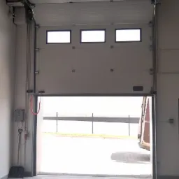 Otwarta brama segmentowa z trzema okienkami w górnej części, widok z wnętrza hali na zewnątrz, w tle zaparkowany pojazd.