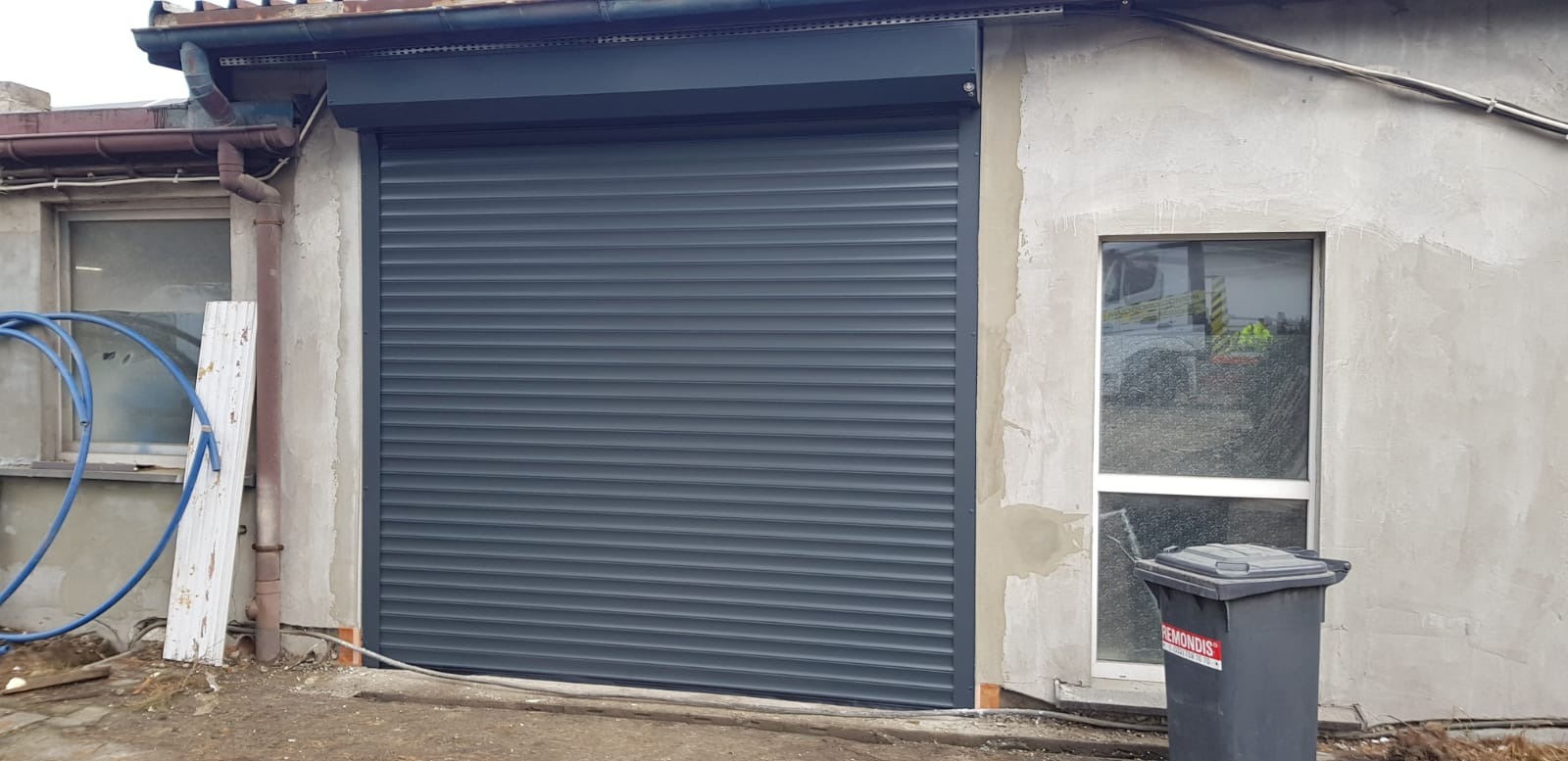 Zamknięta grafitowa roleta zewnętrzna na szarej ścianie budynku z widocznym oknem i rynną.