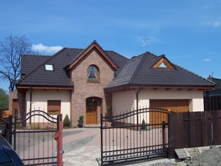 Nowoczesny dom jednorodzinny w Tarnowskich Górach z brązową dachówką i beżową elewacją, widoczna brama wjazdowa z metaloplastyki oraz częściowo zasłonięta brama garażowa z lamelami.