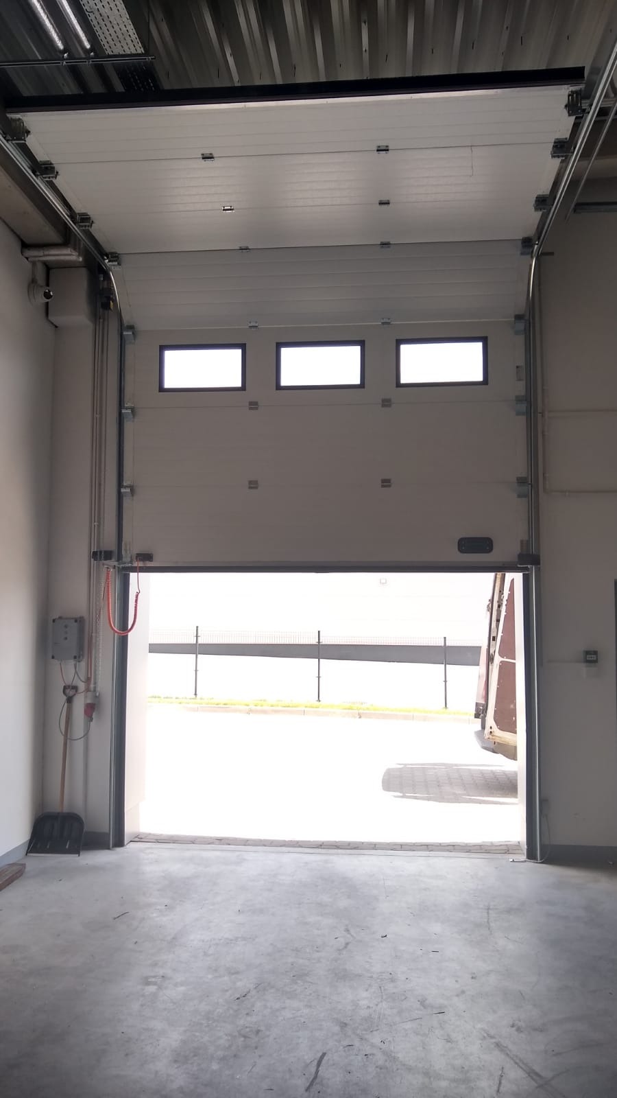 Otwarta brama segmentowa z trzema okienkami w górnej części, widok z wnętrza hali na zewnątrz, w tle zaparkowany pojazd.