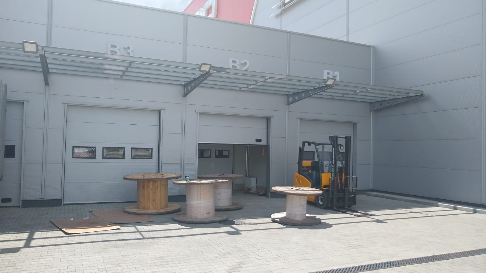 Hala przemysłowa z trzema segmentowymi bramami w kolorze szarym, z numeracją B1, B2, B3, z podjazdem z kostki brukowej i wózkiem widłowym obok otwartej bramy.