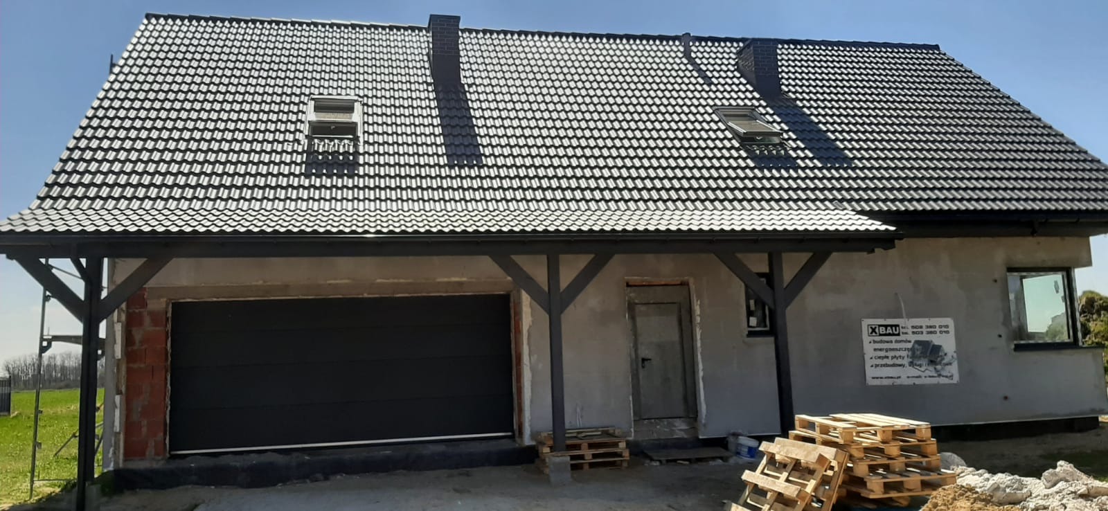 Nowoczesny dom jednorodzinny z grafitową bramą garażową segmentową, dachem pokrytym ciemną dachówką i drewnianą konstrukcją zadaszenia nad wejściem, widoczne palety budowlane.