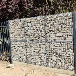 Gabionowe ogrodzenie z metalowymi słupkami, wypełnione jasnymi kamieniami, widoczne na tle drzewa o ciemnych liściach, na nier&oacute;wnym, piaszczystym podłożu.