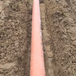 Pomarańczowa rura drenażowa ułożona w wykopie w ziemi, widok z góry.