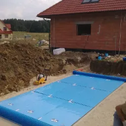 Fundament domu jednorodzinnego w trakcie izolacji termicznej, widoczna niebieska warstwa izolacyjna z rurami drenażowymi, ubijarka obok wykopu.