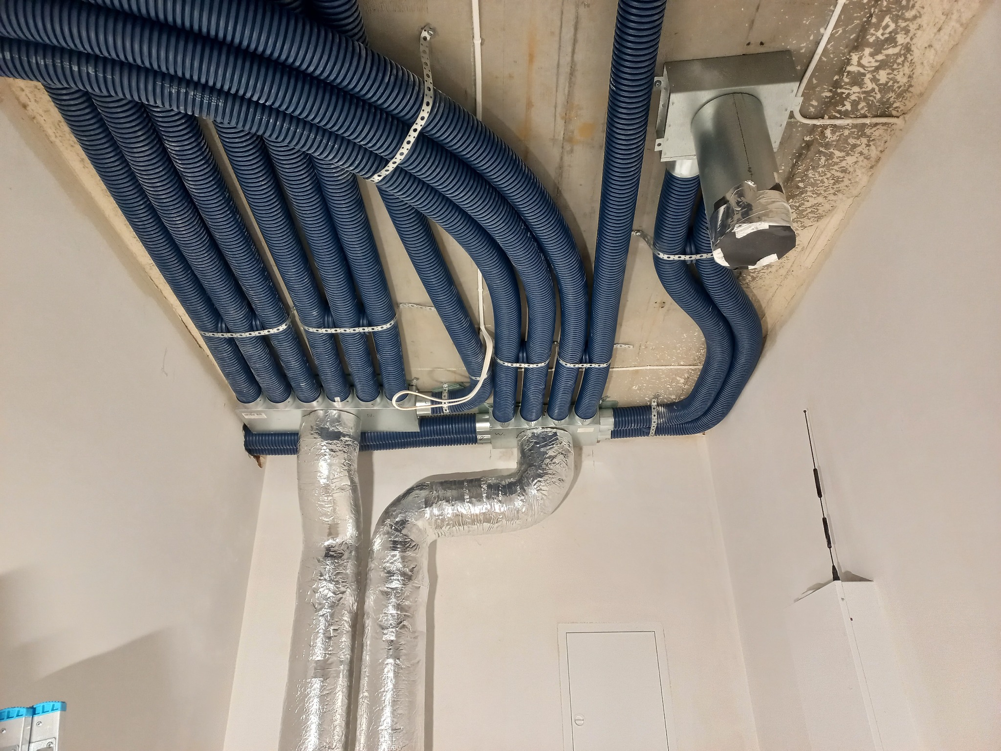 Niebieskie, karbowane rury wentylacyjne pod sufitem, przymocowane metalowymi obejmami. Widoczne srebrne rury izolowane folią aluminiową i biała ściana.