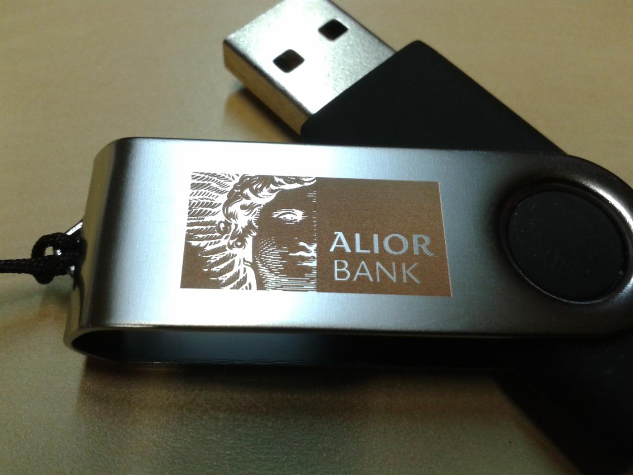 Metalowy pendrive z logo Alior Banku i wizerunkiem klasycznej rzeźby, leżący na beżowym tle.