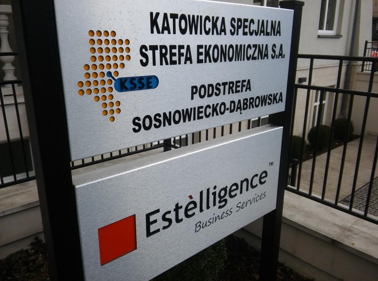 Tablica informacyjna z logo Katowickiej Specjalnej Strefy Ekonomicznej i firmy Estelligence Business Services przed budynkiem.