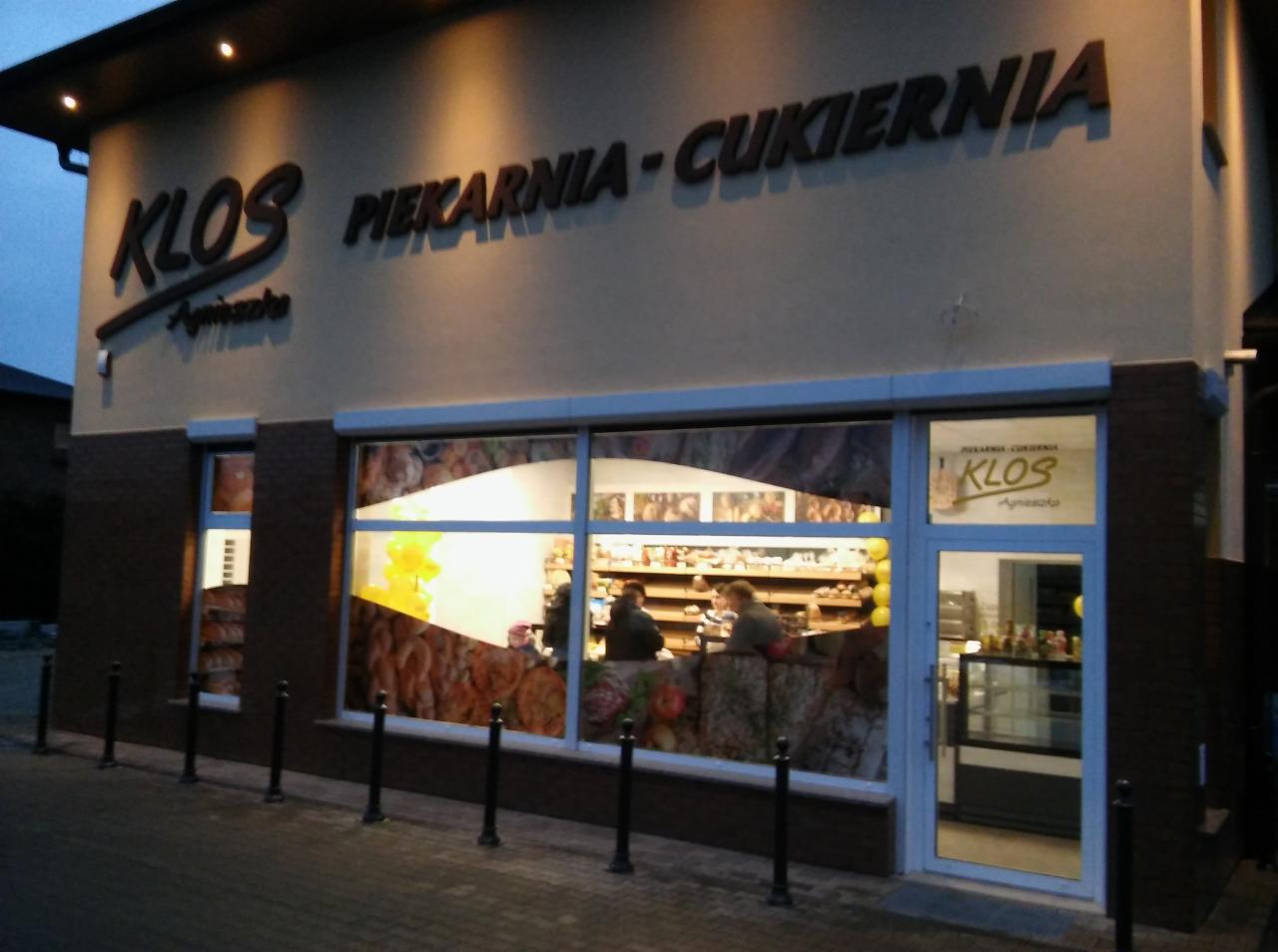 Fasada piekarni-cukierni Klos z dużym oknem wystawowym, na którym widoczne są dekoracje i klienci w środku. Nad oknem umieszczony jest szyld z nazwą firmy.