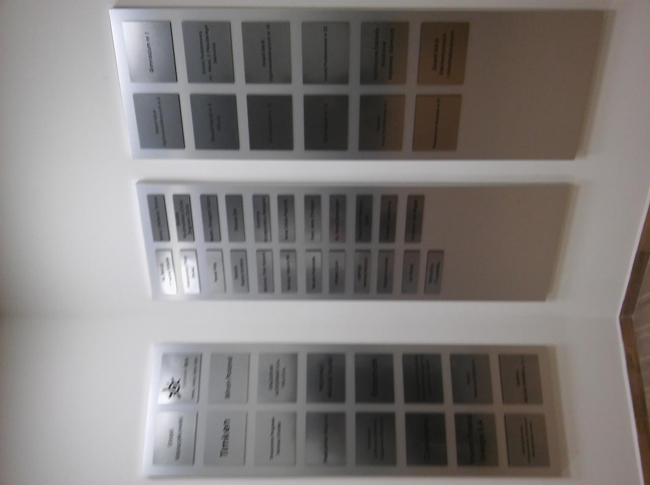 Tablice informacyjne z metalowymi plakietkami z grawerowanymi napisami, zamontowane na białej ścianie.