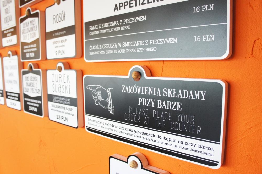 Różne tabliczki z menu i informacją o składaniu zamówień, wiszące na pomarańczowej ścianie w restauracji, wykonane w stylu retro.