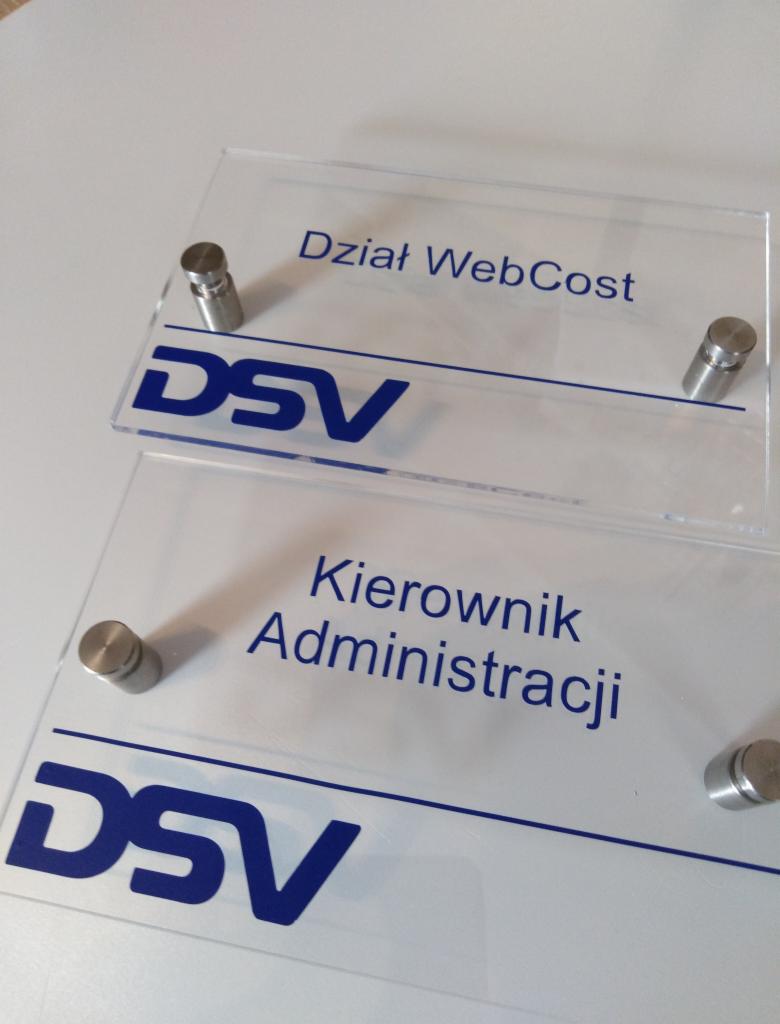 Dwa przezroczyste szyldy z logo DSV, jeden z napisem 'Dział WebCost' i drugi 'Kierownik Administracji', zamocowane za pomocą metalowych śrub dystansowych.