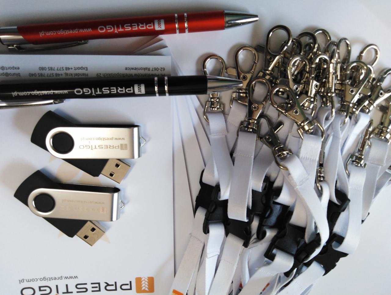 Zestaw promocyjny z logiem firmy: długopisy w kolorze czarnym i czerwonym, pendrive oraz smycze z karabińczykami na białym tle.
