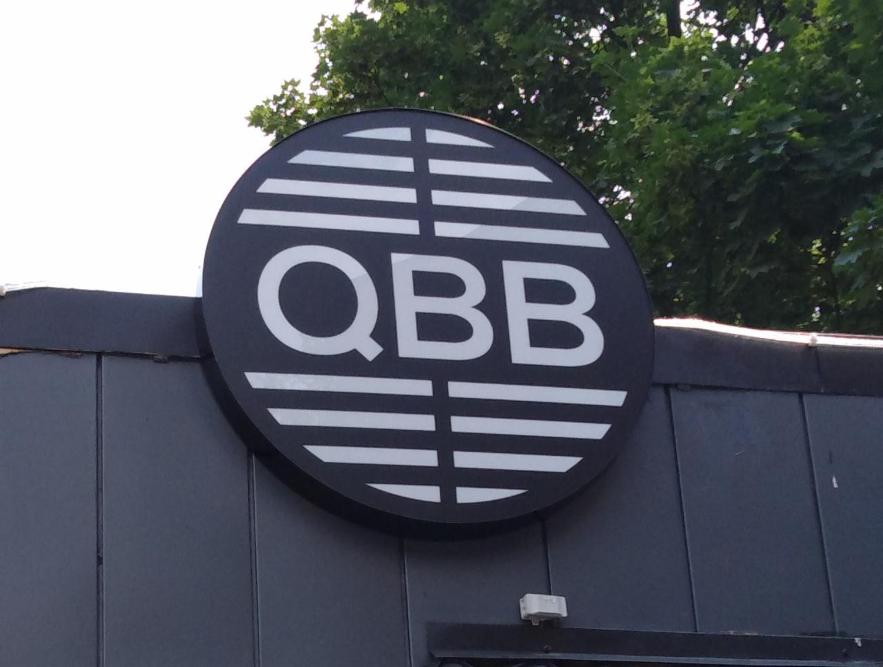 Okrągły szyld z napisem QBB w kolorze białym na czarnym tle, umieszczony na szarej elewacji budynku.