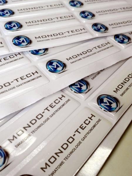 Stos białych, błyszczących etykiet z logo i nazwą firmy 'MONDO-TECH' oraz tekstem 'ŚWIATOWE TECHNOLOGIE GASTRONOMICZNE'.